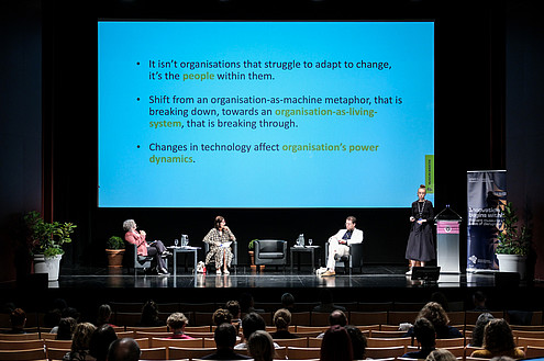 Three people are sitting on a stage. Another person is standing and talking to an audience. In the background, presentation slides are projected to a screen.   © Image: Jorge Gomes