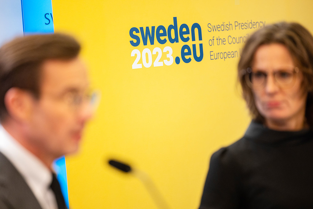 © Image: Axel Öberg/Regeringskansliet This photograph shows two people in the foreground who are out of focus. The background consists of a yellow wall and the logo of the Swedish Presidency of the Council of the European Union.
