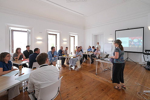 People are sitting on small tables in a conference room. One person is standing and talking. Presentation slides are projected to a screen at the front of the room.  © Image: Jorge Gomes