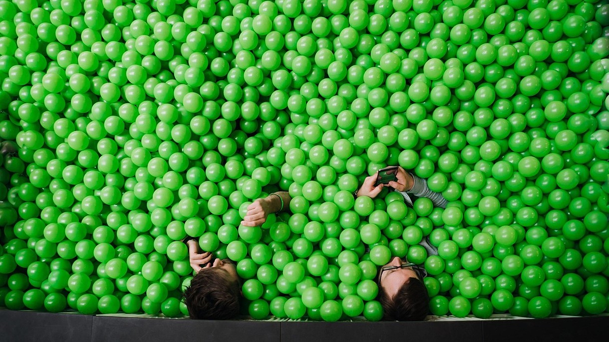 © Who Am I / Alamy Stock Foto Two people. photographed from bird perspective, sit in a pit of green plastic balls. They are both using their smartphones.