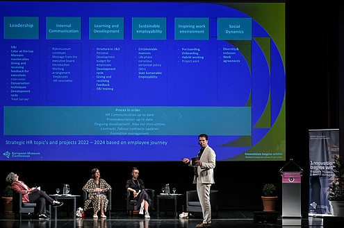 Three people are sitting on a stage. Another person is standing, gesturing with their arms and talking. In the background, presentation slides are projected to a screen.   © Image: Jorge Gomes