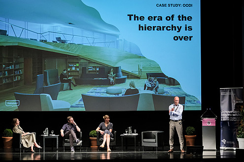Three people are sitting on a stage. Another person is standing with their arms crossed and talks. In the background, presentation slides are projected to a screen.   © Image: Jorge Gomes