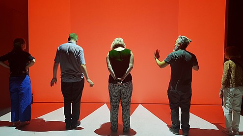 Five people with their back to the camera are standing in front of a big orange wall.  