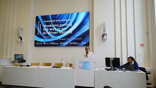  A person is standing behind a podium talking into a handheld microphone. Behind her there is a projected image with the name of the conference "Innovation and Integrity: Museums paving the way in an AI-driven society".