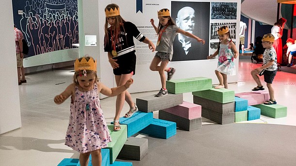  Children with paper crowns on their heads are balancing on stones inside an exhibition space.