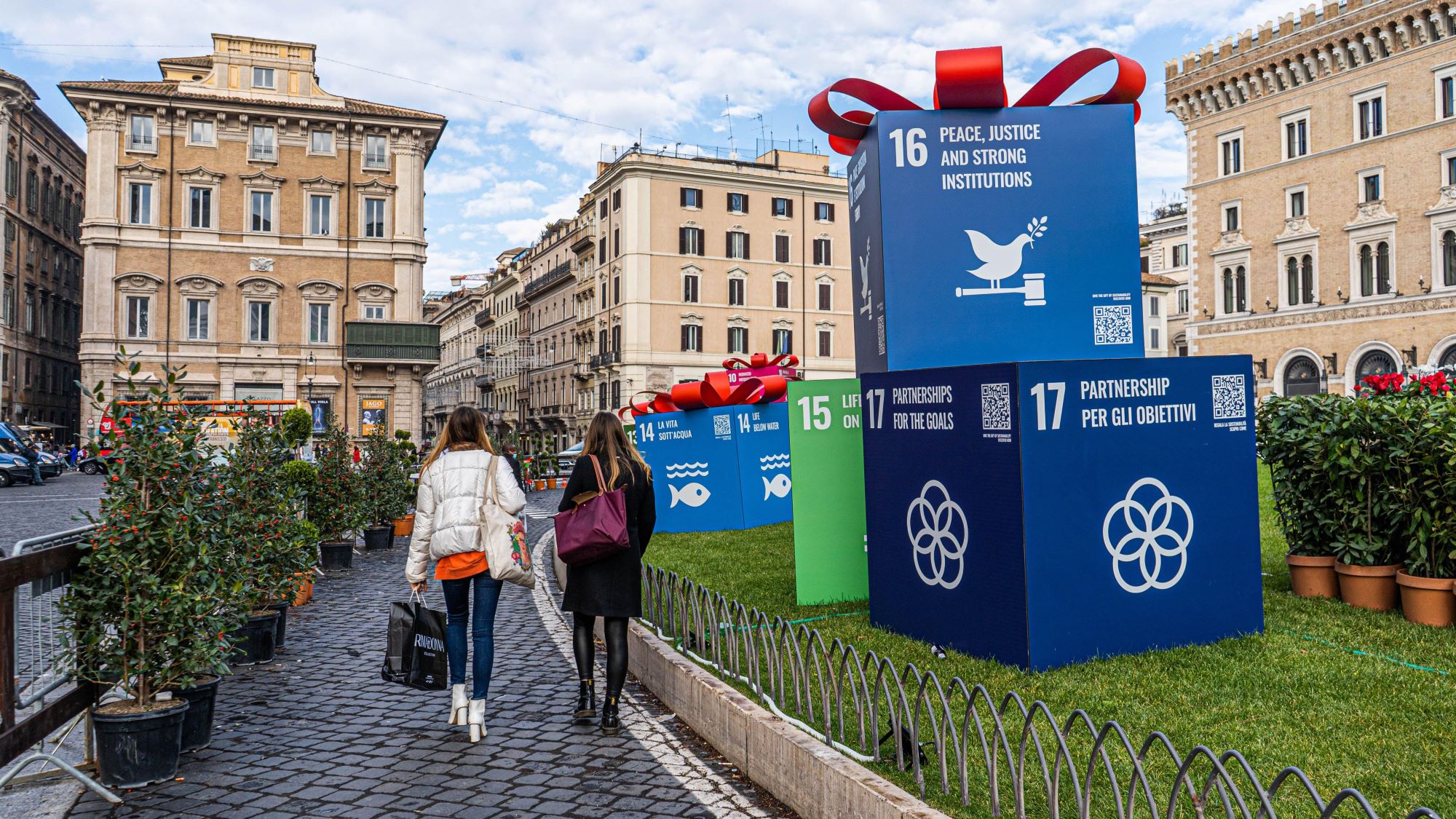 Two people are walking across a town square that is decorated with big boxes representing the UN's Sustainable Development Goals. Readable are boxes with goal 16 Peace, Justice and strong institutions and goal 17 Partnerships for the goals.  © Alamy Stock Photo, Image: Amer Ghazzal