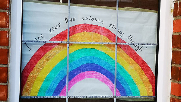 The photographs shows the window of a brick house. Behind the window a poster is attached, which shows a rainbow and reads "I see your true colours shining through".