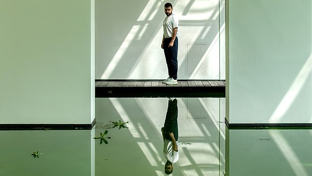  A person is standing on a footbridge inside a room with a floor covered by a mirror. The person is looking at their reflection.