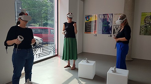Three people standing in a gallery space wearing VR glasses and holding VR joysticks.  