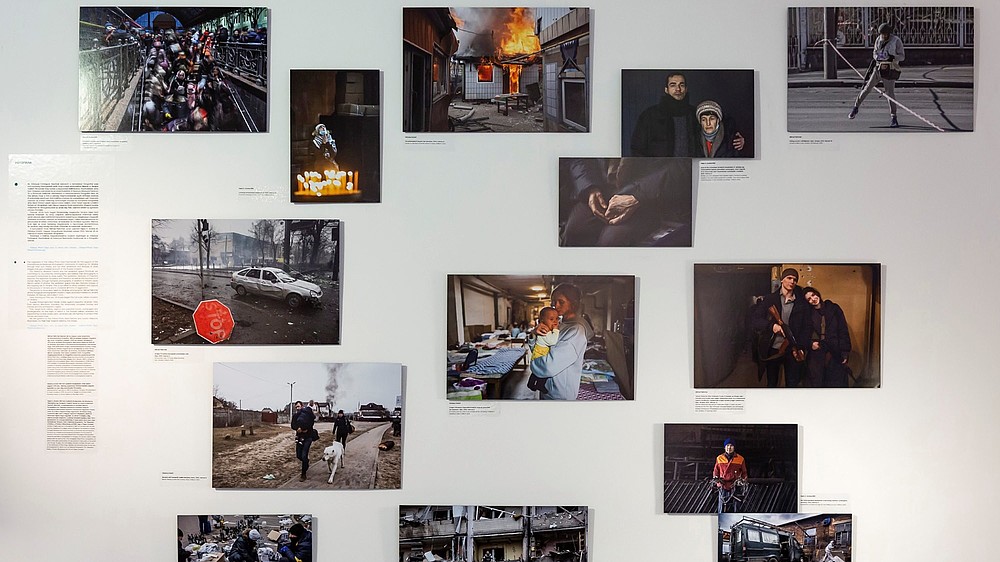 Various photographs are mounted on to a wall with accompanying text. The photographs show the war in Ukraine.  