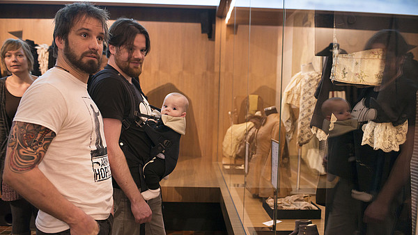 Two men stand next to each other in front of a glass monter displaying old fashioned clothes. One of the men is carrying a baby in a harness on his belly.
