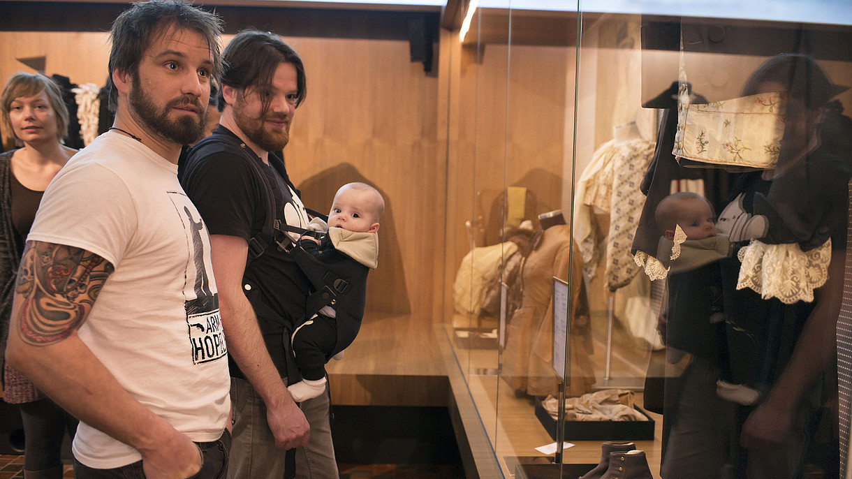 © Zeeuws Museum, Image: Anda van Riet Two men stand next to each other in front of a glass monter displaying old fashioned clothes. One of the men is carrying a baby in a harness on his belly.