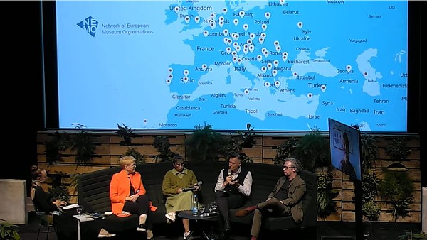 Five people, photographed from above, sit on a couch on a stage in a dimly lit room. One of them talks and holds a microphone. On the wall a map of Europe is projected.