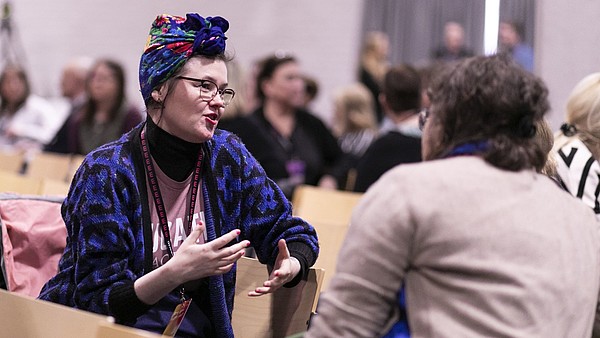 Two people are talking. One of them has their back to us, the other one is dressed in bright colours, facing the camera and is gesticulating with her hands while explaining something.