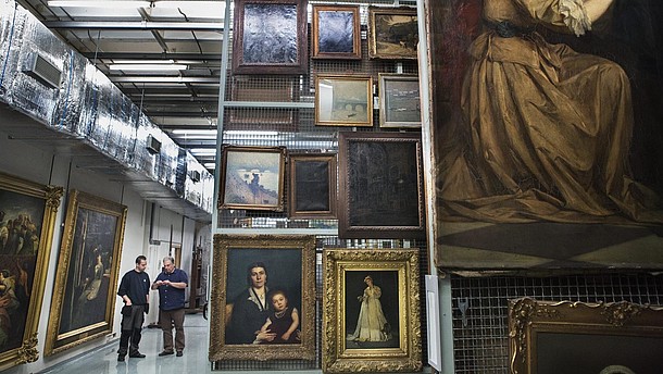  Two people are walking through a museum's depot filled with paintings.