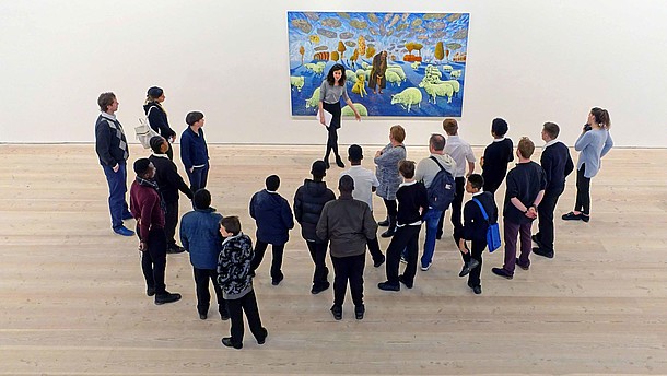  A group of people stand in front of a modern painting together with a museum educator. The image is photographed from bird perspective.