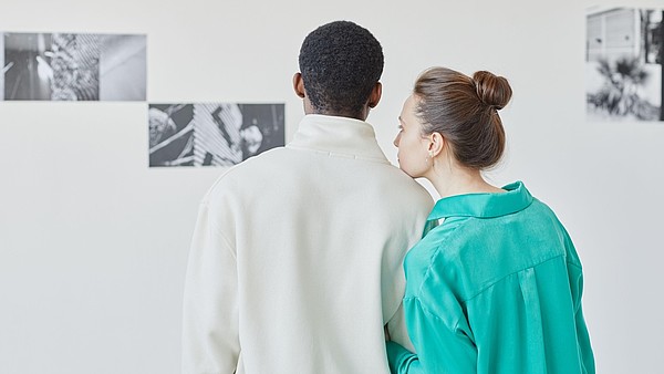 Two people are standing in a gallery looking at black and white photographs on the wall. One of them is resting their head on the others shoulder.