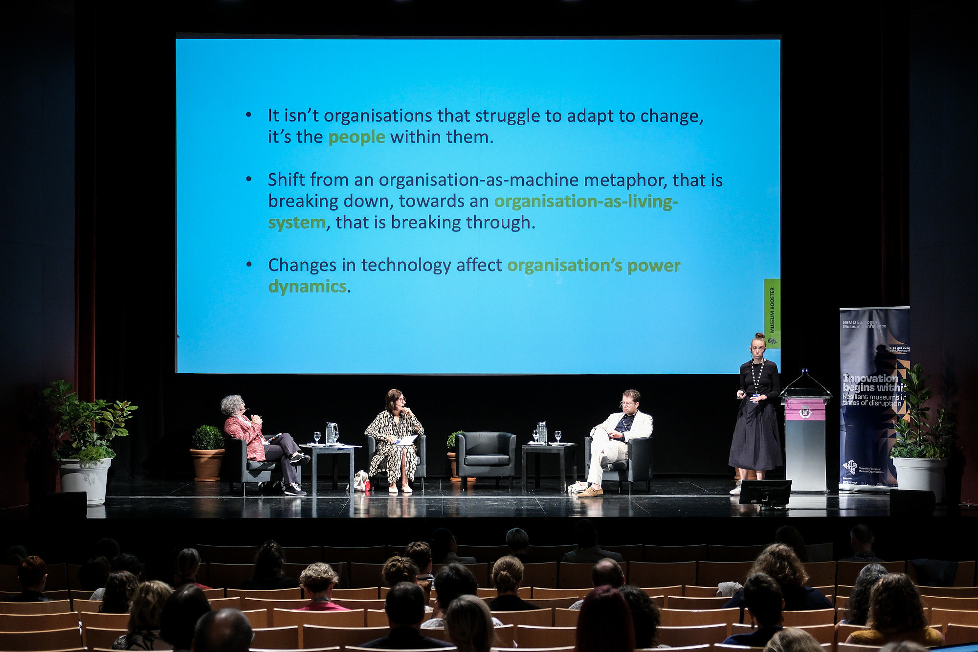 Three people are sitting on a stage. Another person is standing and talking to an audience. In the background, presentation slides are projected to a screen.   © Image: Jorge Gomes