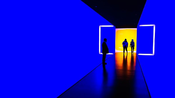 Three people walk through a light installation. They walk from a room lit in yellow into a blue tunnel. 
