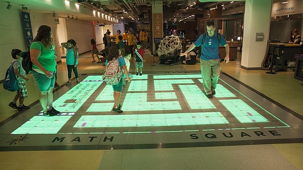 A group of children and adults interact with a digital projection on the floor.