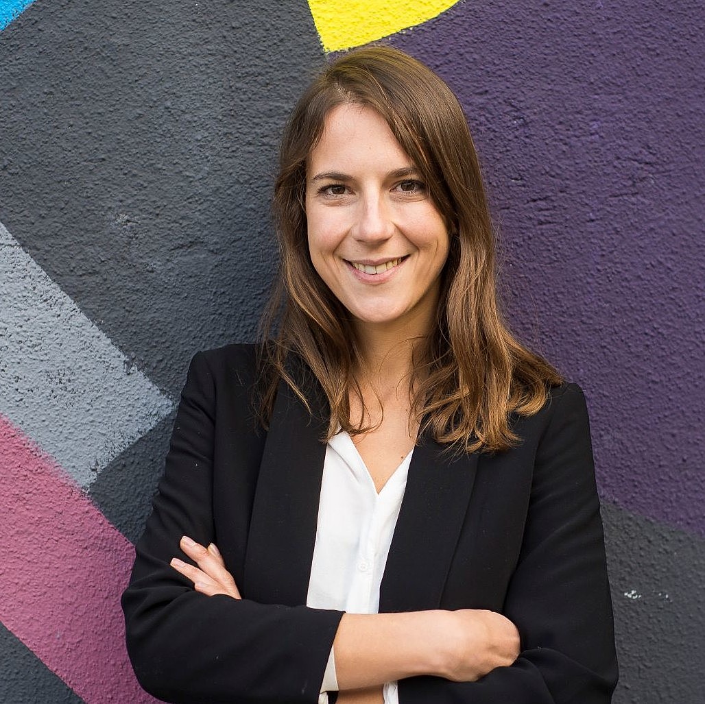  Portrait of a person leaning against a colourful wall with their arms crossed and smiling into the camera.
