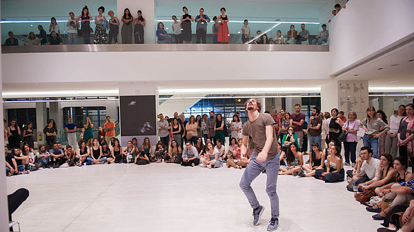 A person is dancing, surrounded by a number of people sitting or standing and watching them. 