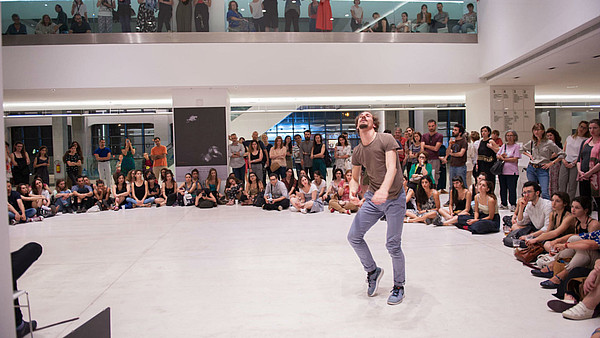 A person is dancing, surrounded by a number of people sitting or standing and watching them. 
