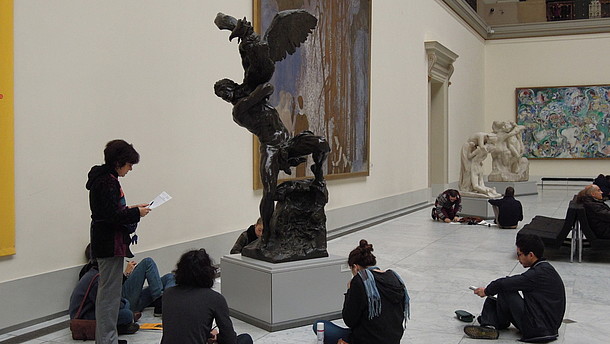  A group of young people is sitting around a sculpture in a museum space. Another person is standing and holding a piece of paper.