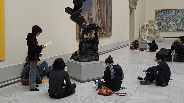 A group of young people is sitting around a sculpture in a museum space. Another person is standing and holding a piece of paper.