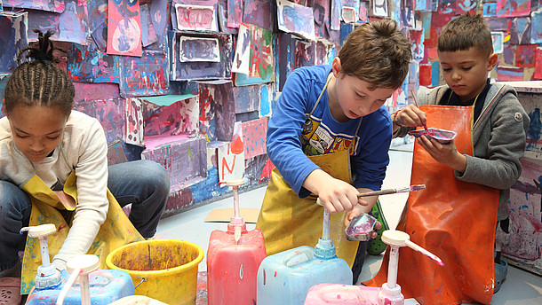  Three children are mixing paint colours. The wall behind them is covered with colourful paintings and cardboard boxes.