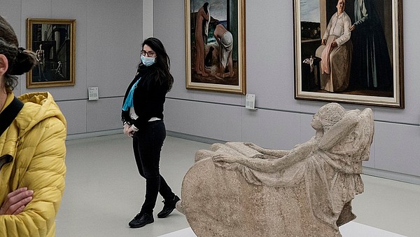 A museum guide is walks through an exhibition room filled with paintings and a stone sculpture. Another person is partly seen on the left side.