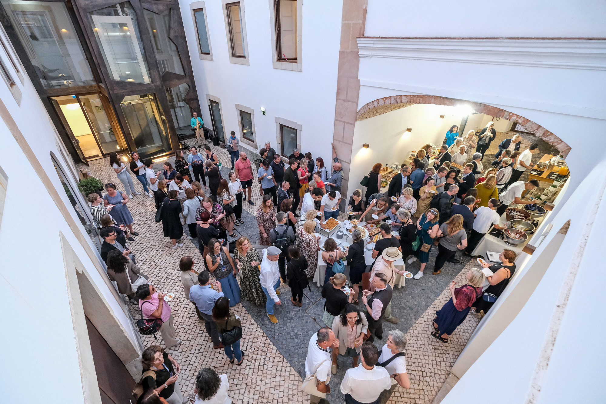 Photo taken from bird perspective of a large group of people standing inside a courtyard. Some people are standing by a buffet, others are socialising or eating.  © Image: Jorge Gomes
