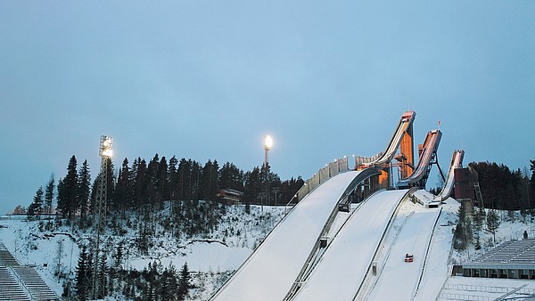 Three ski jump towers covered in snow. Next to the towers is a snowy hill with pine trees.
