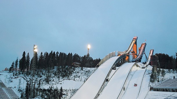  Three ski jump towers covered in snow. Next to the towers is a snowy hill with pine trees.