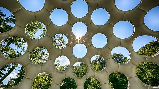 View upwards and through a roof structure with big circular holes. Behind blue skies and trees are visible.