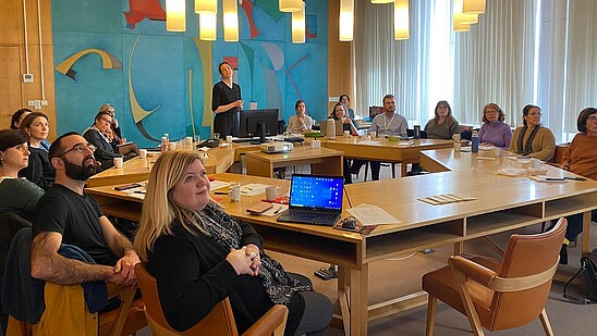 This photograph depicts various people sitting around a hexagon-shaped table. They are looking up in the same direction. One person stands and is talking to them.