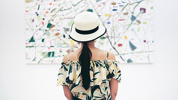 A woman wearing a hat is standing with her back to us. She is in centre of the picture and of the painting in front of her.