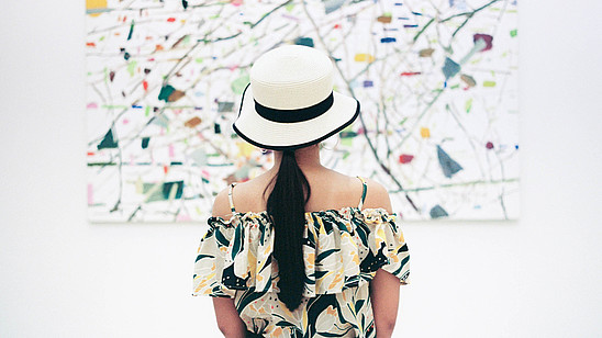 A woman wearing a hat is standing with her back to us. She is in centre of the picture and of the painting in front of her.