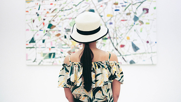  A woman wearing a hat is standing with her back to us. She is in centre of the picture and of the painting in front of her.