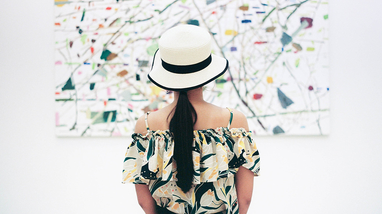 © Image: Vincent Tantardini A woman wearing a hat is standing with her back to us. She is in centre of the picture and of the painting in front of her.