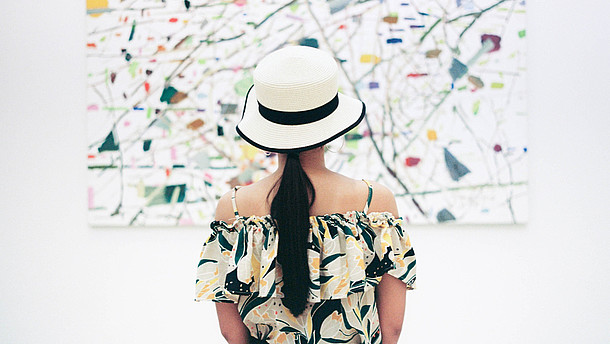  A woman wearing a hat is standing with her back to us. She is in centre of the picture and of the painting in front of her.