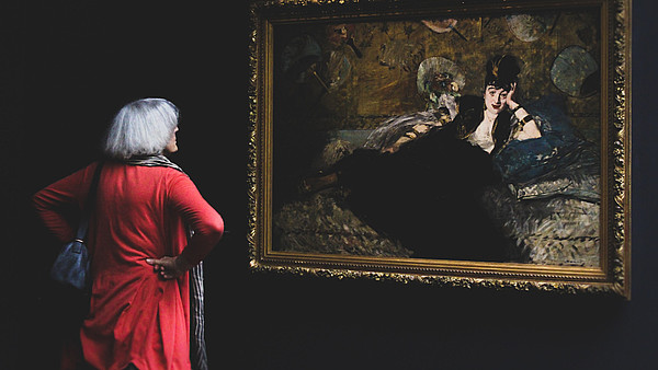 Woman with her back to the camera is looking at a painting of a person in a black dress. The wall is also black which contrasts the woman's red dress.