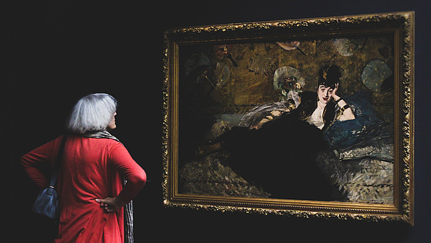  Woman with her back to the camera is looking at a painting of a person in a black dress. The wall is also black which contrasts the woman's red dress.