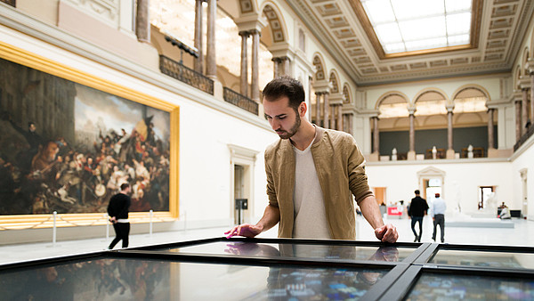 A man is looking down on an art piece that isn't visible in the photo. He is standing in a big bright gallery space with columns and vaults. There is a huge painting on one of the walls and people in the background.