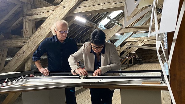 Sabine Verheyen leans forward to cut a paper with a big paper cutter board. A person is standing to her side to assist. 