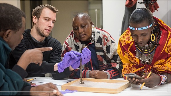 A museum professional wearing gloves is holding an object and presenting it to a group of three people. Two are wearing colourful traditional clothing. One is looking attentively at the object, while the other is looking at their phone. The third person is photographes from the back indicating at the object.
