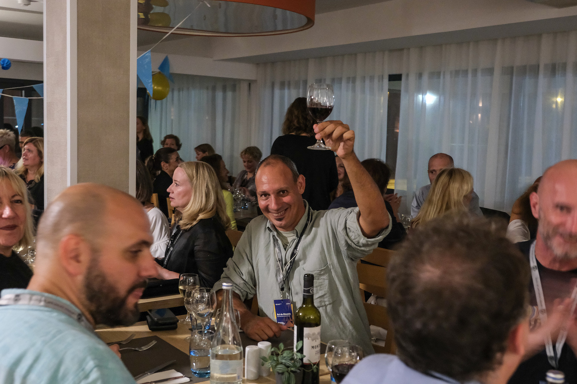 People are sitting at a restaurant table and talk to each other. In the middle of the picture one person is raising their glass and looks directly at the camera.   © Image: Jorge Gomes