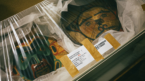  This photo shows two ancient art objects packaged inside a box with foil on top. On the foil is a sticker of the "Humboldt Forum" as well as an inventory sticker.