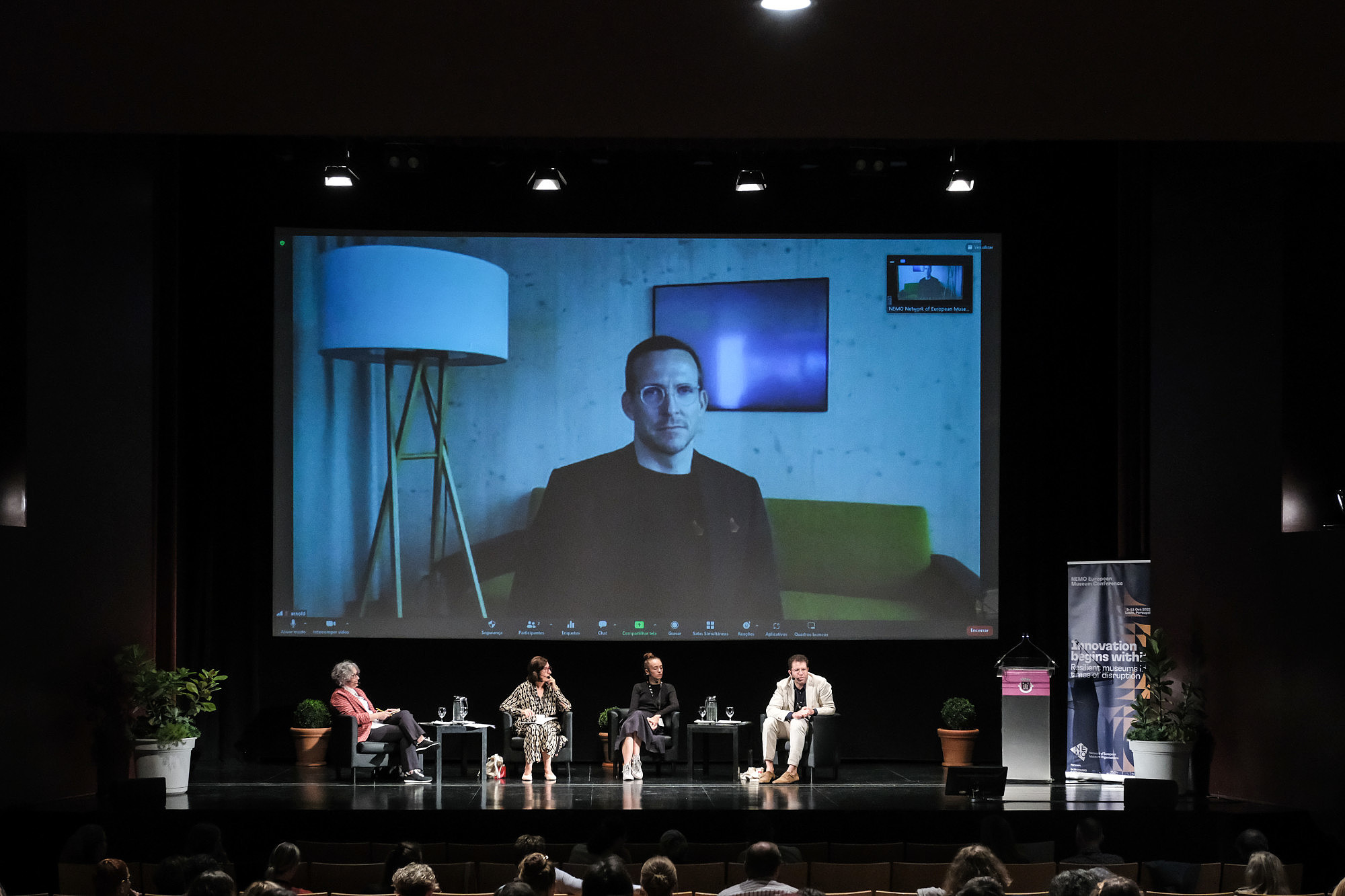 Four people are sitting on a stage and look towards an audience. A person in a video call is projected to a screen behind them.   © Image: Jorge Gomes