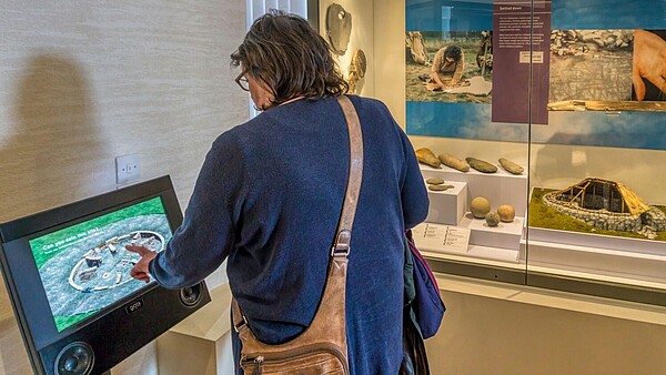 A museum visitor stands in front of a showcase with ancient objects and uses a touchscreen standing next to it.